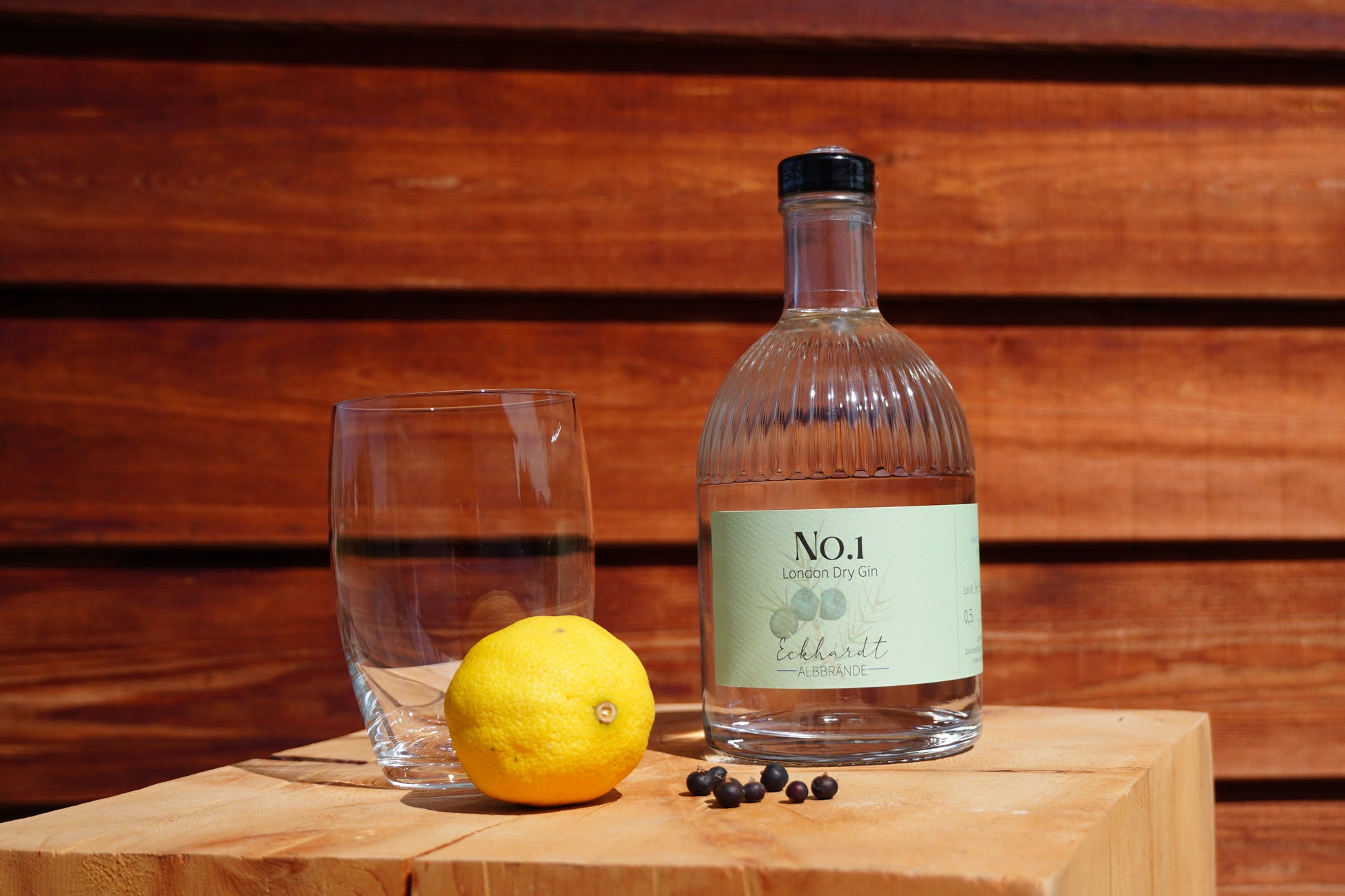 Flasche mit London Dry Gin No.1 von Eckhardt Albbrände auf einem Holzblock vor einer hölzernen Wand. Daneben steht ein leeres Glas, außerdem liegt auf dem Holzblock eine Zitrone und Wacholderbeeren.