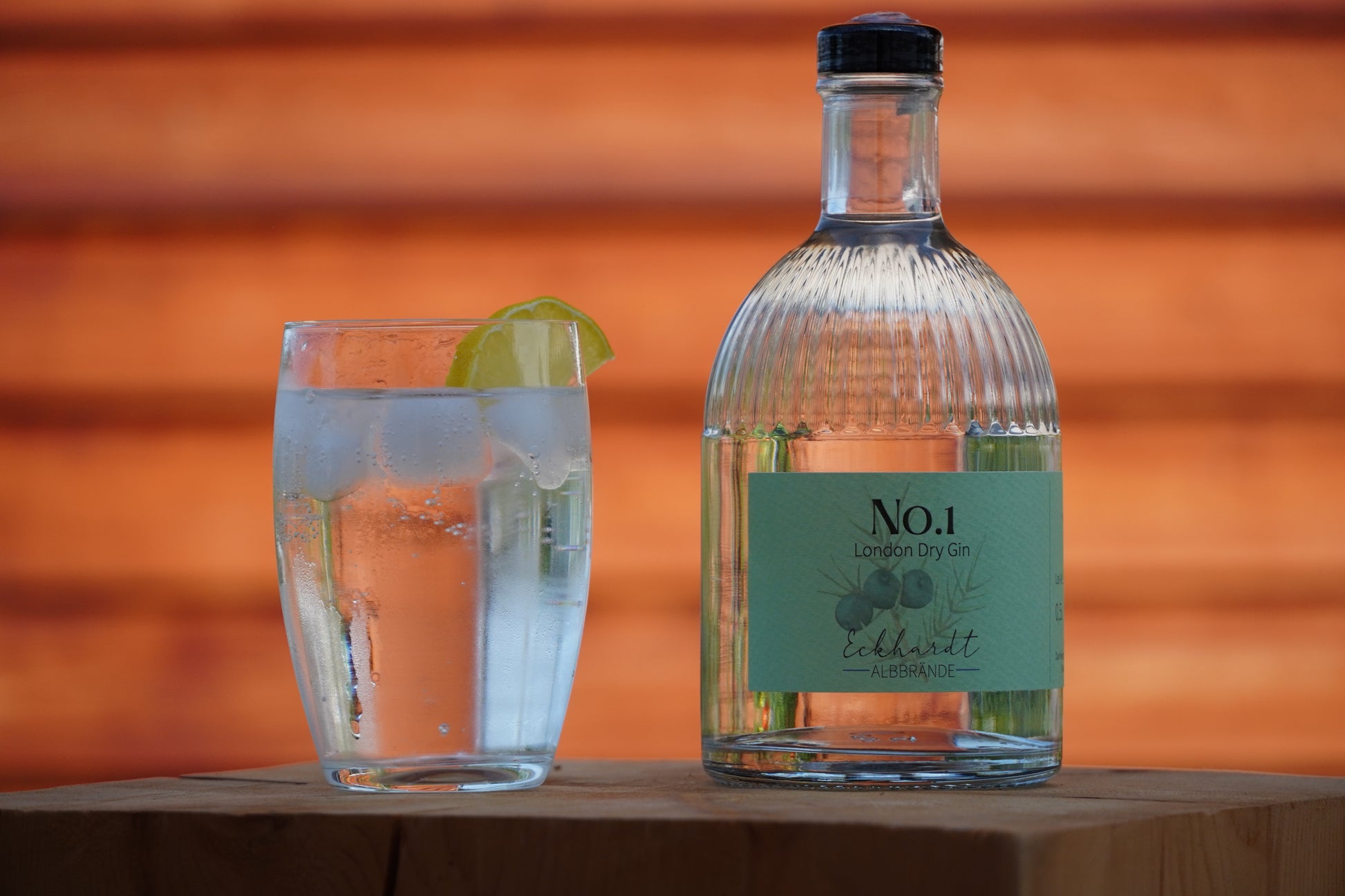 Flasche mit London Dry Gin No.1 von Eckhardt Albbrände auf einem Holzblock. Daneben steht ein Glas mit Gin Tonic, Eiswürfeln und einer Zitronenscheibe.
