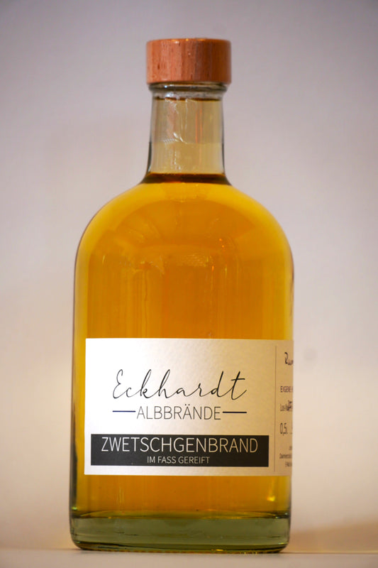 Glasflasche mit Holzdeckel und eine braun-orangefarbenem Inhalt vor einem weißen Hintergrund. Auf dem eleganten Etikett steht der Schriftzug "Eckhardt Albbrände" und die Bezeichnung "Zwetschgenbrand im Fass gereift".